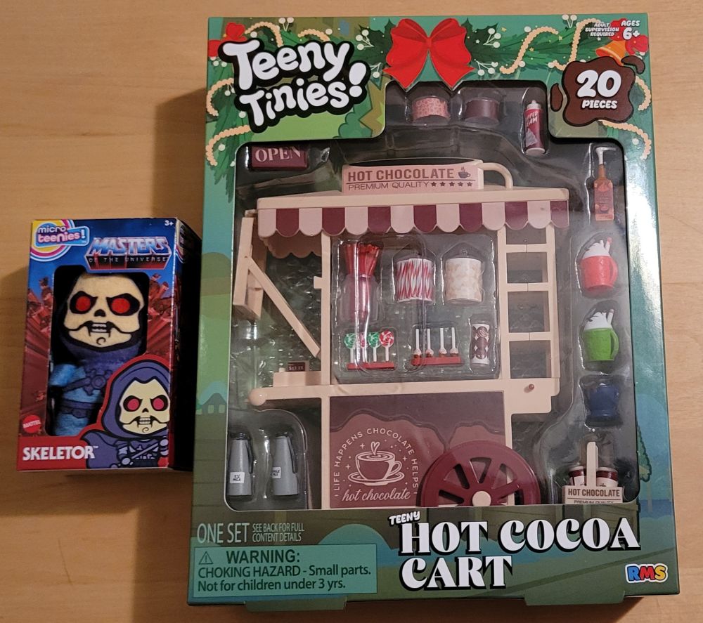 A small plush Skeletor in a cardboard Masters of the Universe box and a set of Teeny Tinies- Hot Cocoa Cart. The cart is beige and has about 20 accessories, including some mugs, toppings, and candy, among other things. The box is green and Christmas themed. 
