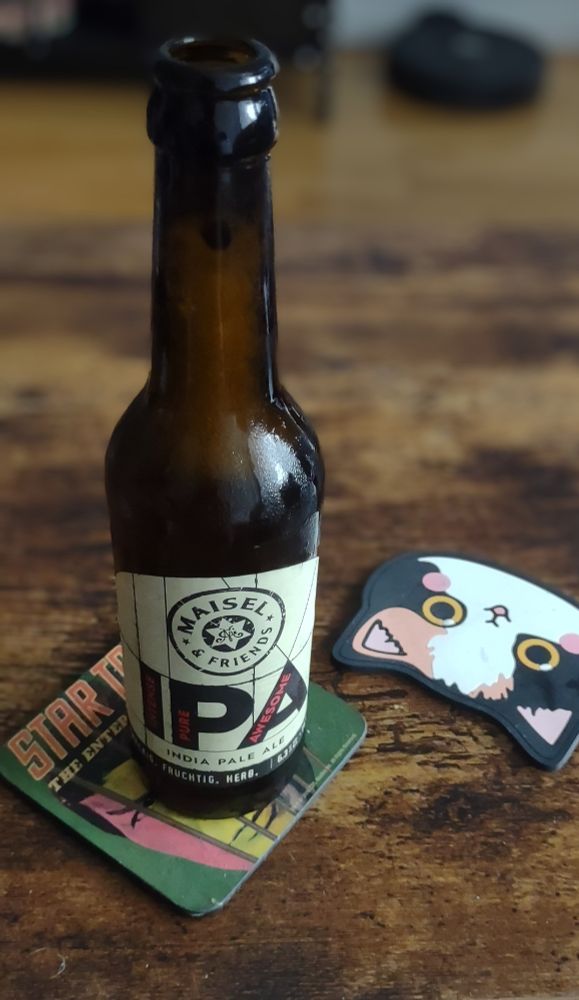 Garrafa de cerveja long. No rótulo bege se le a palavra IPA em letras garrafais. Está sob um coaster onde se lê parcialmente Star Trek. Há um segundo coaster ao lado em formato de cara cartunizada de gato. Tudo sobre uma mesa de madeira marrom escuro. Ao fundo se vê um rack de tv desfocado