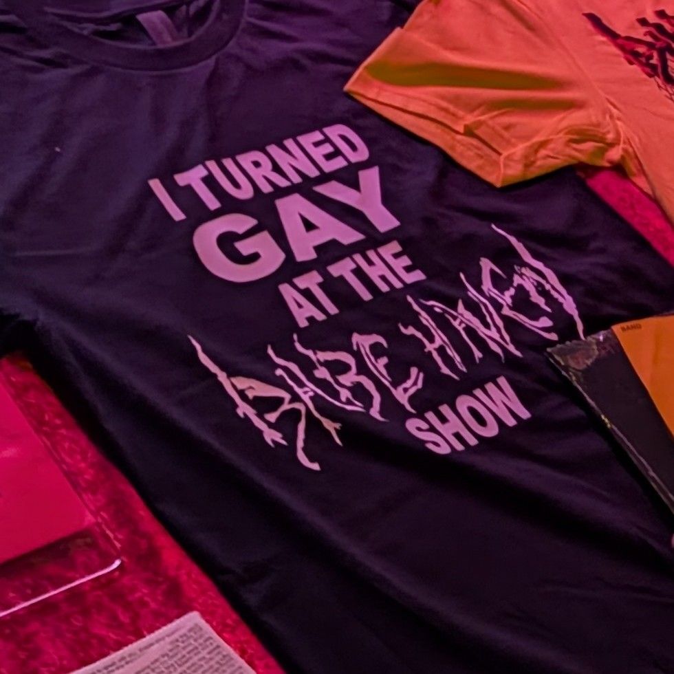 Merch table with black T-shirt that says "I TURNED GAY at the BABE HAVEN Show"