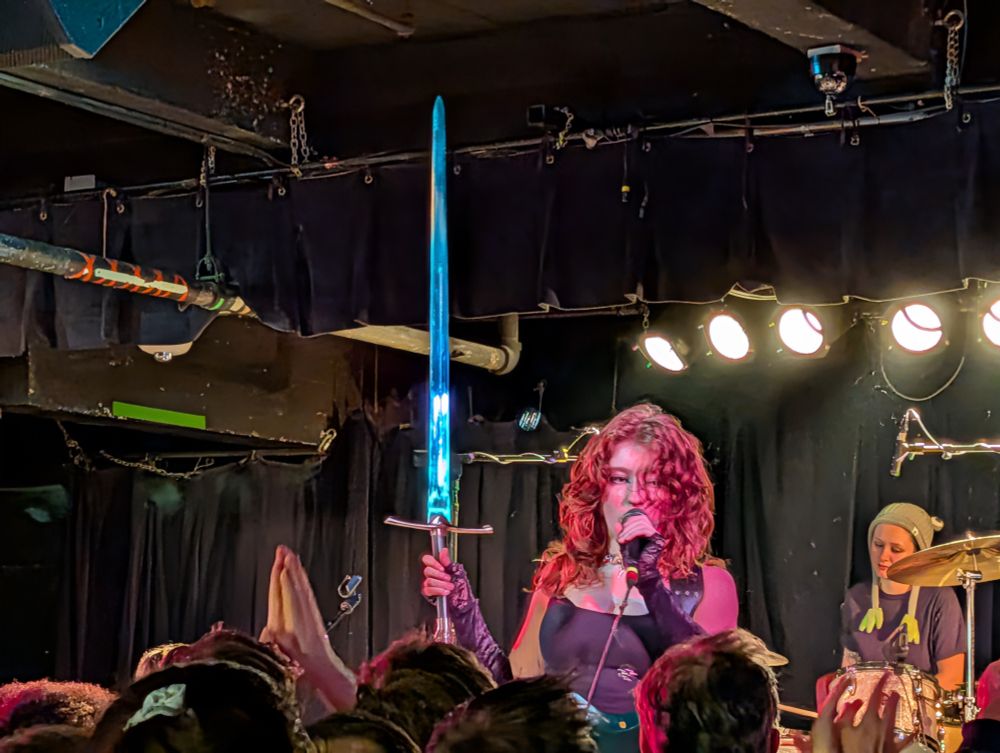 A person with curly red hair, wearing a black top, holds a glowing blue sword high above their head while singing into a microphone on a dimly lit stage in front of a crowd. A drummer is visible to the right.
