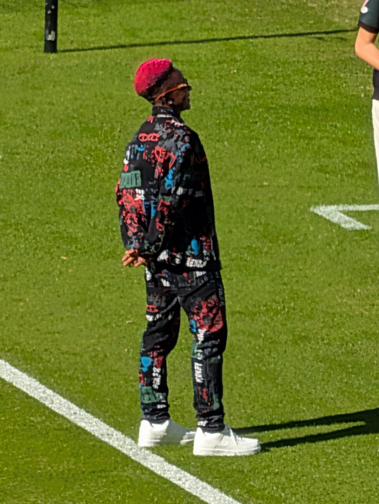 A person with bright pink hair wearing a black jacket and matching pants with colorful graffiti-style graphics, standing on a green grass sports field.
