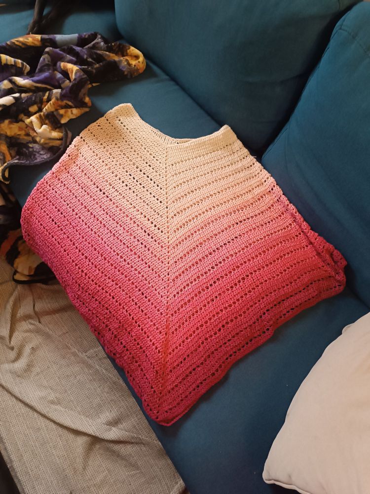 Photo d'un poncho léger fait au crochet posé sur un canapé bleu. Le poncho est dans un dégradé qui va du blanc au rose foncé. 