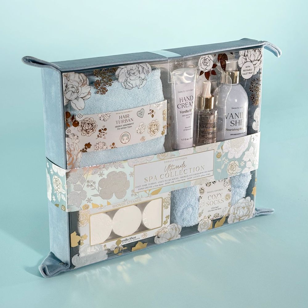 In front of a light blue backdrop is an aqua suede tray, filled with a set of bath and body items by Beauty Concepts. Items include a hair turban, plush socks, and vanilla shea shower steamers, hand cream, body oil and body lotion. The entire set is decorated with silver and gold rose designs.