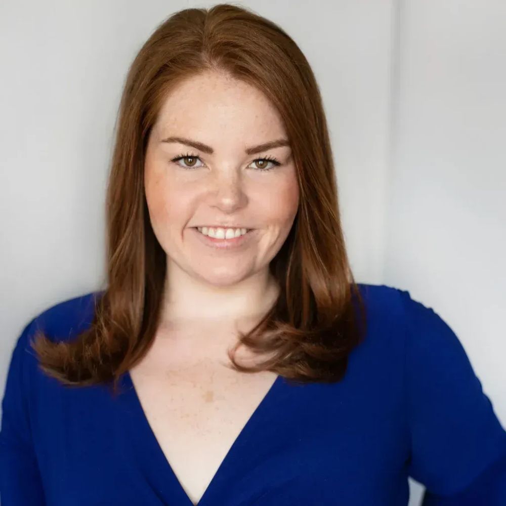 Headshot of Sky, a white freckled person with medium length red hair and brown eyes. They're wearing a blue dress and smiling