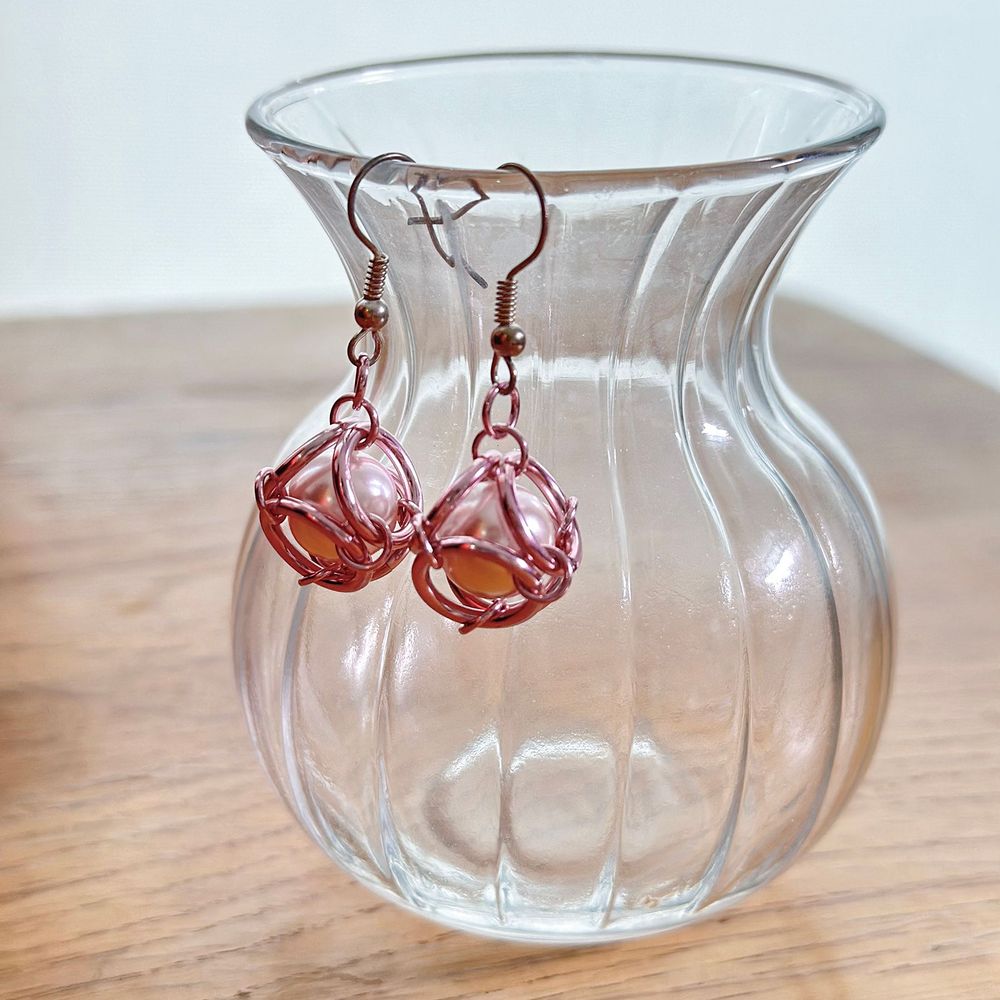 Closeup of Artemis earrings hanging from a glass vase, featuring pearl drops caged within interlocked pink chainmail rings.