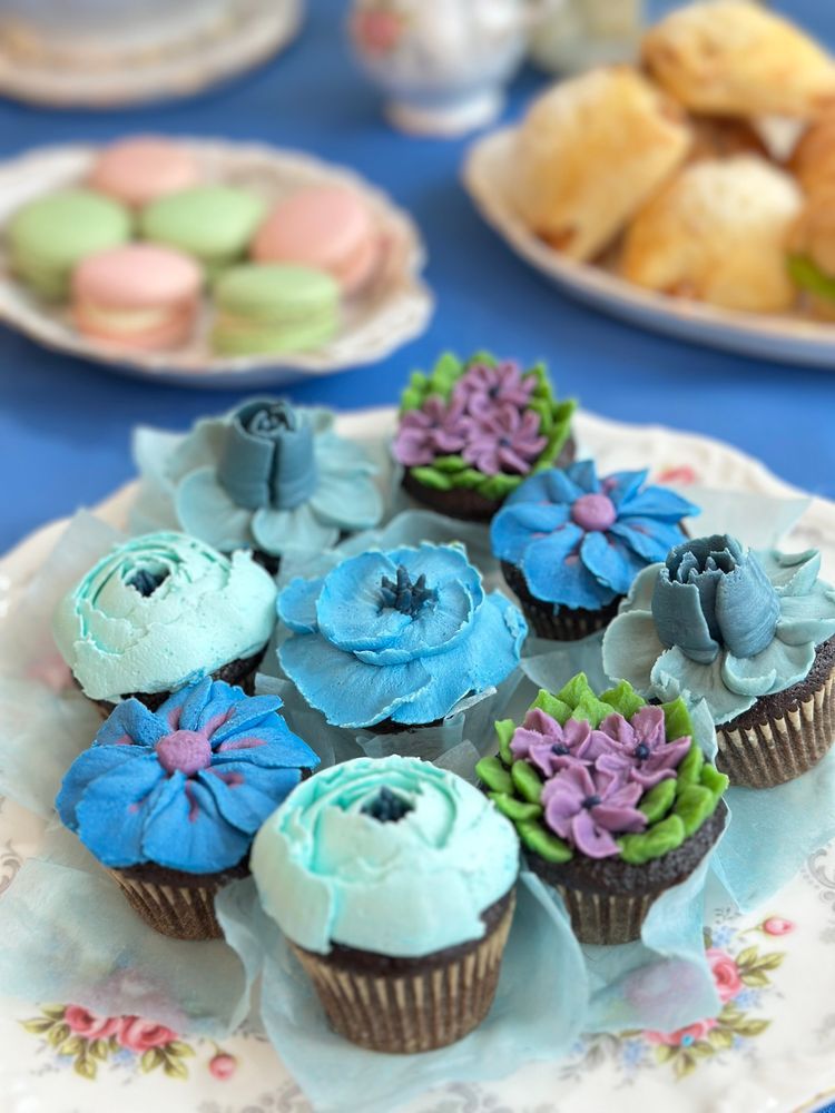 Closeup of nine buttercream-frosted chocolate cupcakes from Maya's Cupcake Bouquets. They're decorated to look like elaborate flowers, including a variety of blue roses and dainty clusters of purple blossoms. The cupcakes sit on a porcelain rose-pattern plate, on a blue tablecloth with a faint butterfly pattern. In the background can be glimpsed some out-of-focus scones and a plate of pink and spring green macarons.