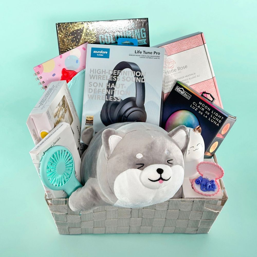 Against an aqua background is a woven grey gift basket containing noise cancelling headphones, a grey and white plushie shaped like a shiba inu, a colouring book, pot-it fidget toy journal, moon light, fairy light photo clips, lavender heat pack, and aqua migraine ice cap.