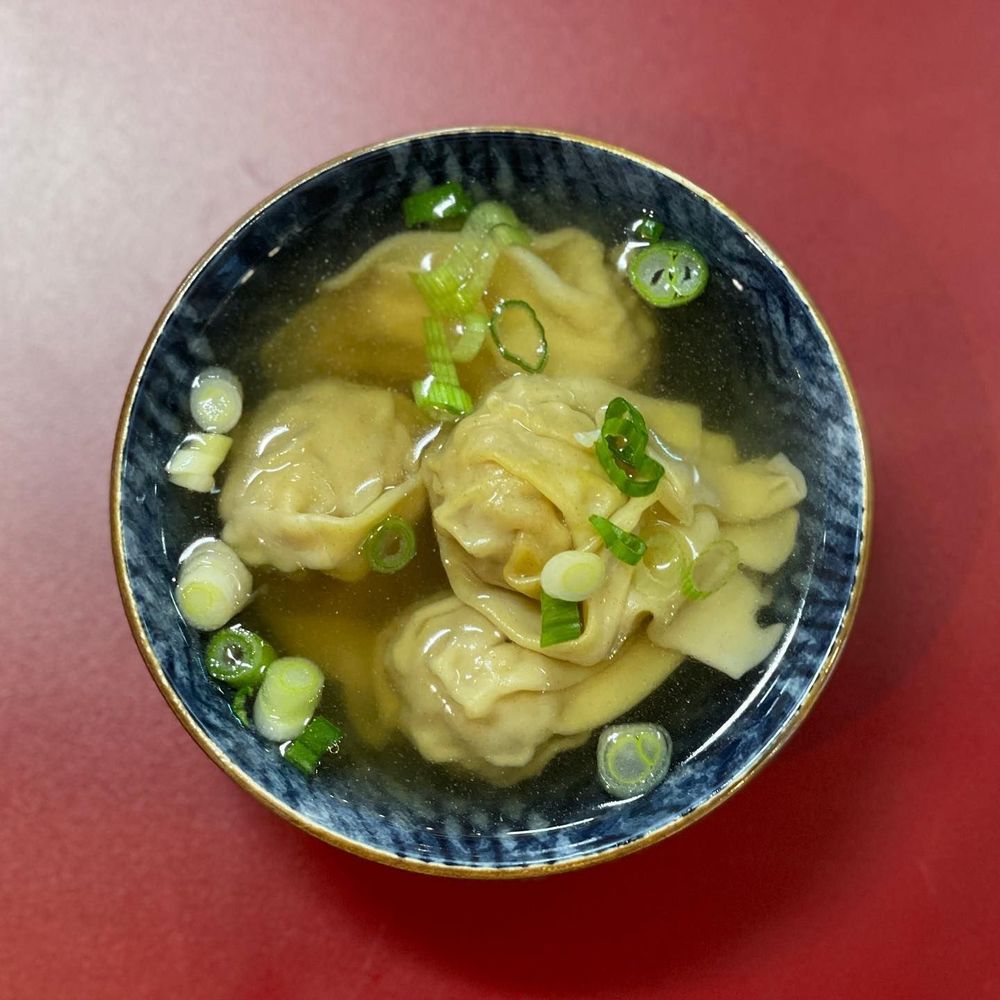 Closeup of wonton dumplings and green onions in broth, in a blue bowl on a red background