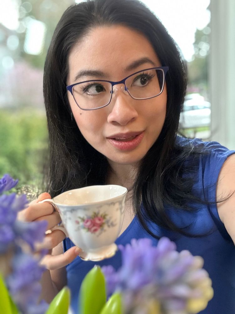 A closeup of Mariposa general manager Stephanie Ko, seen past a screen of out-of-focus blue-purple hyacinth flowers. She is a disabled Chinese-Canadian woman with long dark hair, blue-rimmed glasses and a sea blue top, holding a porcelain teacup with a pink and silver rose pattern, glancing off to the side with a smile on her face. The background behind her is out of focus, with gleams of of light and green foliage glinting through a window.