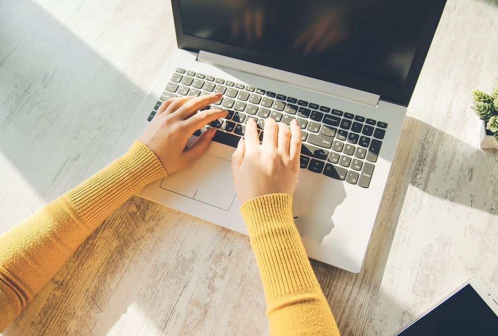 Closeup of a white woman's hands typing on a laptop, her arms clothed in yellow sleeves.