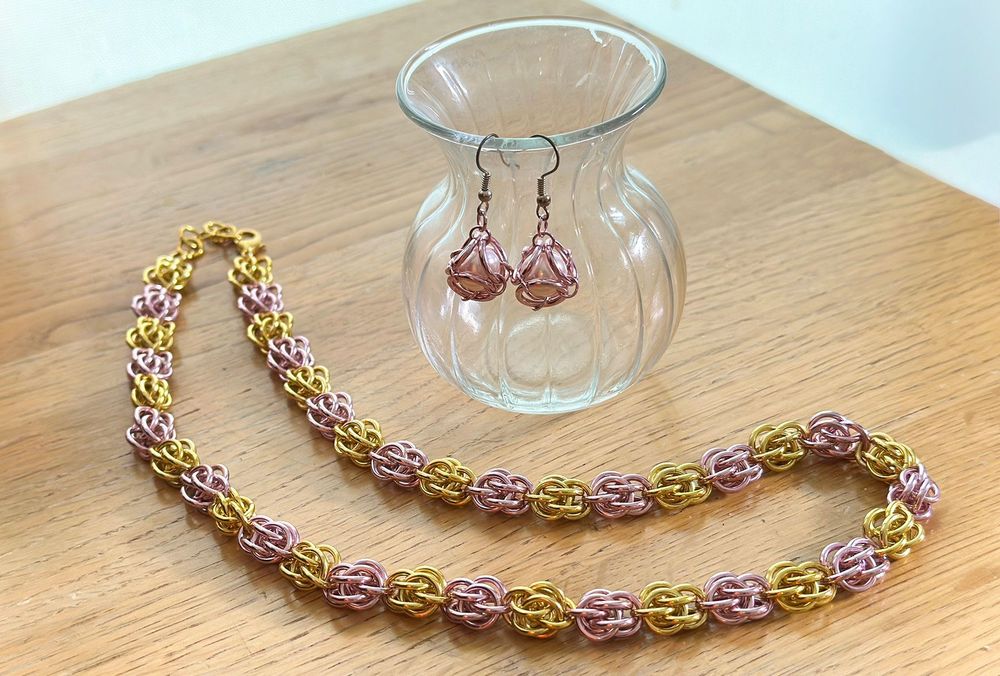 Against a wooden surface, two pearl earring drops are caged in interwoven pink chainmail rings, hanging from a glass vase. In front of that is a necklace that looks like it's made of intricate pink and gold alternating beads, but each bead is woven out of chainmail rings.