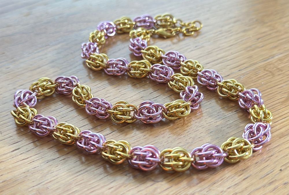 Closeup of the Sweetpea necklace against a wooden surface, featuring alternating pink and gold "beads" woven out of chainmail rings.