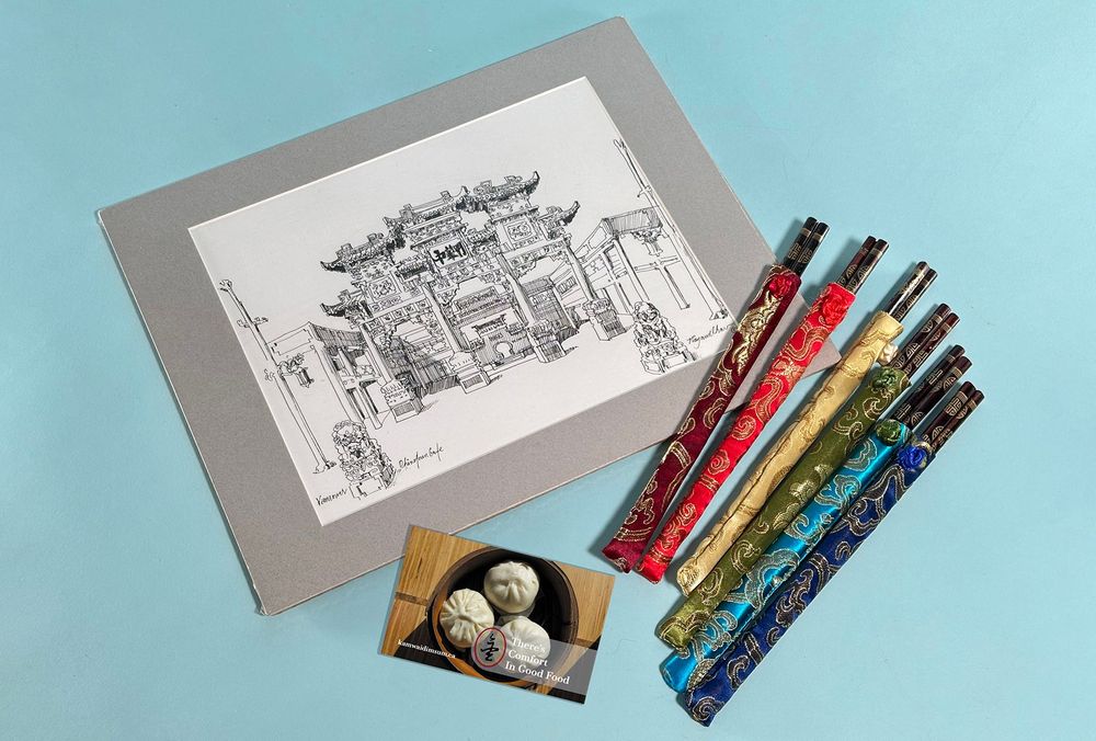 Against a light blue background is a matted print of a Raymond Chow ink drawing of the Chinatown arch. Scattered next to it are a set of six chopsticks wrapped in colourful brocade, and a Kam Wai Dim Sum gift card with an image of pork buns.