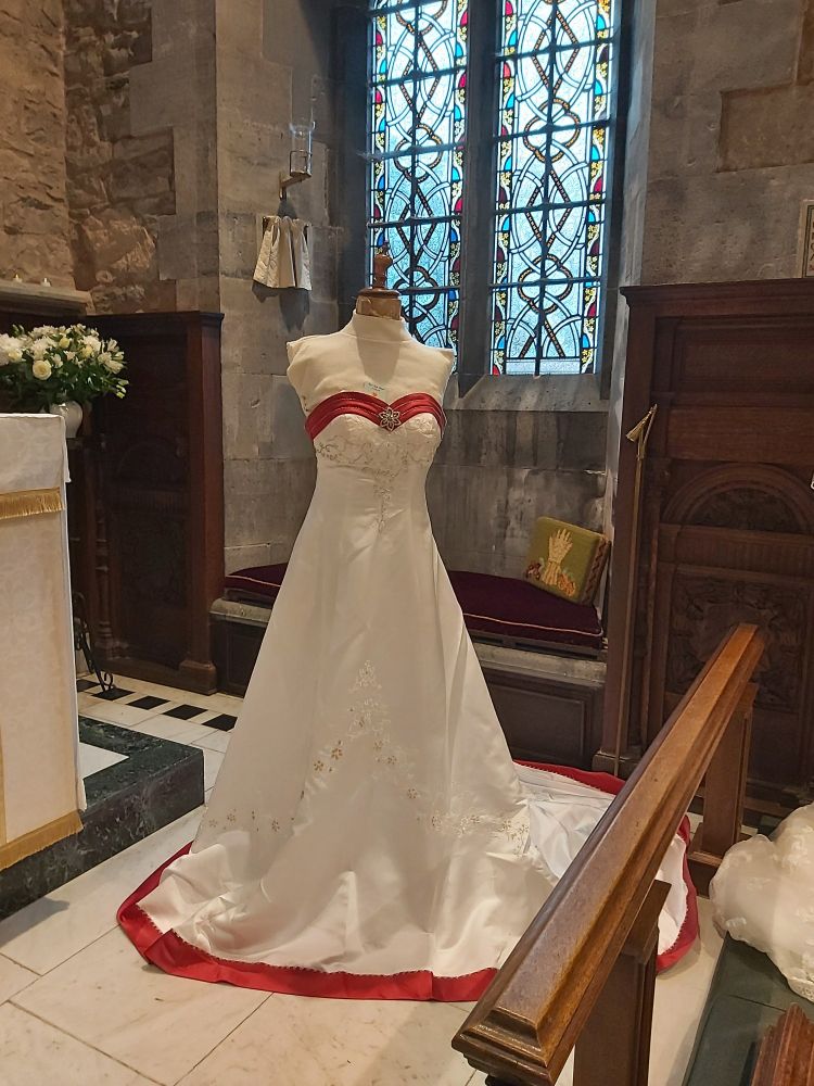 A white sleeveless wedding dress with red trim displayed on a mannequin at the front of the church. Worn by a bride married her 17 years ago.