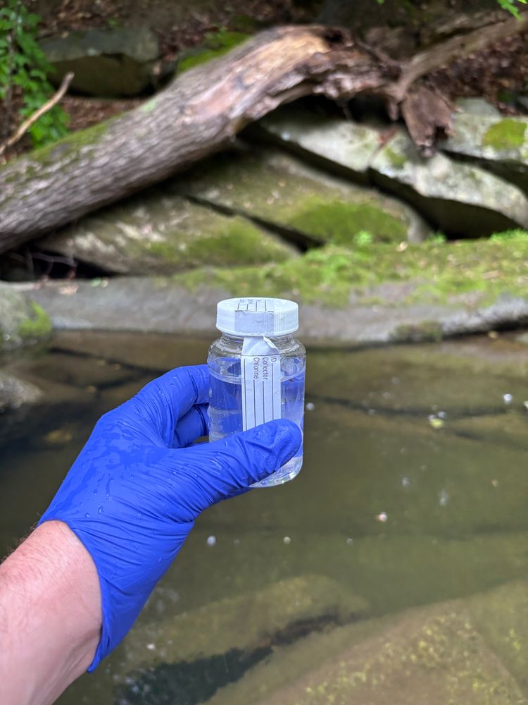 Gloved hand holding a clear plastic sampling bottle filled with water, with stream pool in background