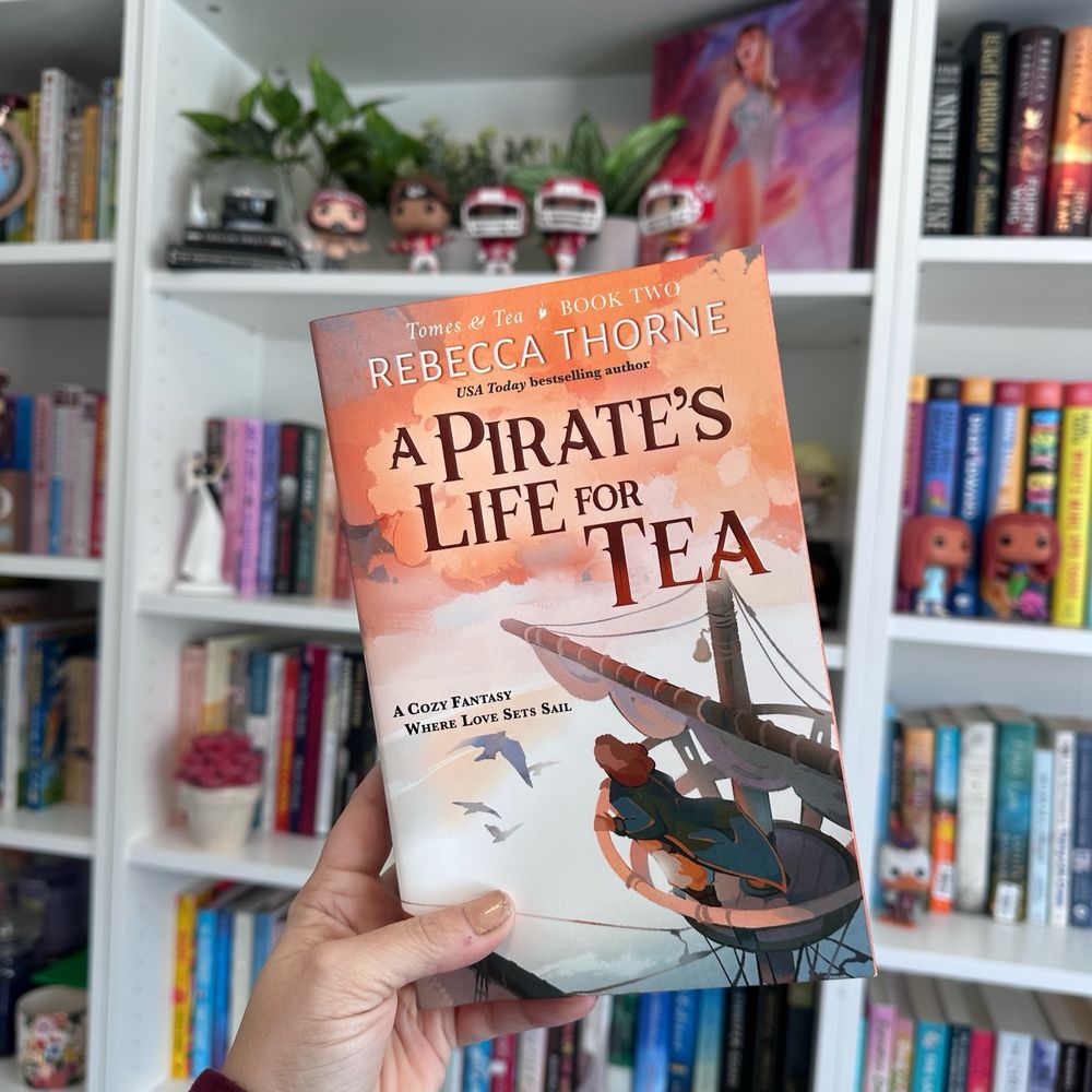 White woman holding up the book, A Pirate’s Life for Tea, in front of bookshelves.