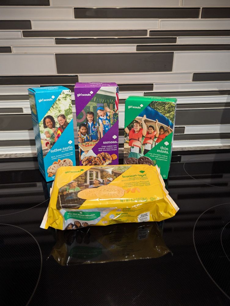 One box each of toffee-tastic, samosa, and thin mint girl scout cookies lined up, with a package of lemon-ups in front of them. A white and black patterned backsplash behind them