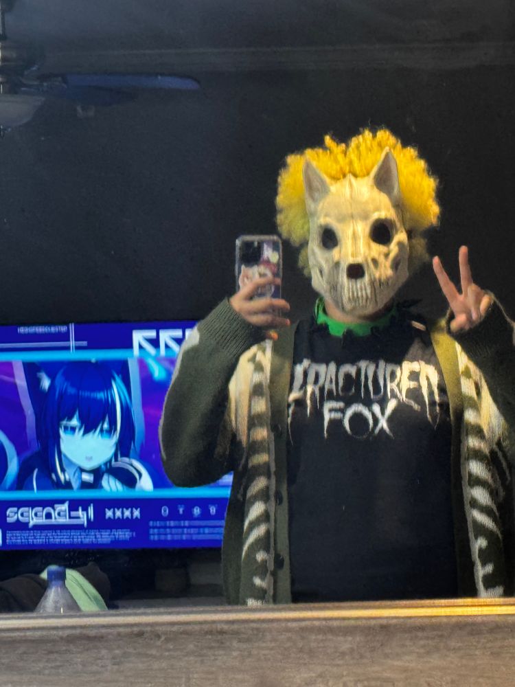 bec is holding a phone and a peace sign, wearing a black shirt with Fracturedfox white distressed text, a dog skull mask, green spiked collar, with a neon green afro. there is a TV in the left background frame with a screenshot of an anime cat girl postermascot for a hyperpop youtube playlist