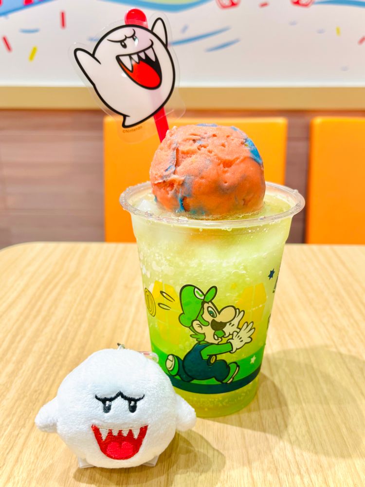 サーティワンのマリオコラボドリンク　ルイージのカップにメロンシロップのかき氷、その上に赤いりんごシャーベット、その上にテレサのピックがついている　手前にテレサの小さいマスコット