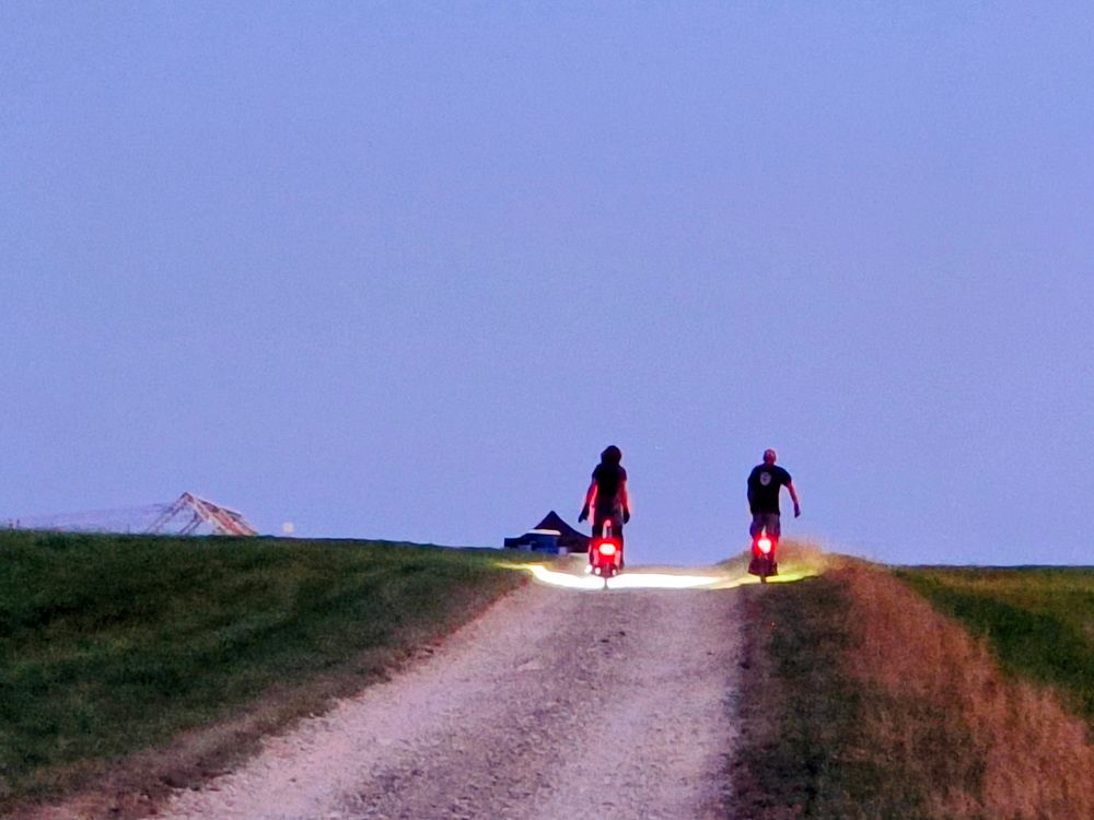 Photo of people on electric unicycles from the back going up a hill on a stony track with red lights

The evening sky is a dim blue

There is a triangular structure in the left corner and the moon can be seen faintly
