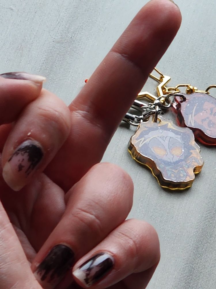 A decrepit photo of a phone charm depicting the Machine Herald from League of Legends and a mildly blurry hand flipping him off. Jayce (Giopara) is slightly out of frame. 