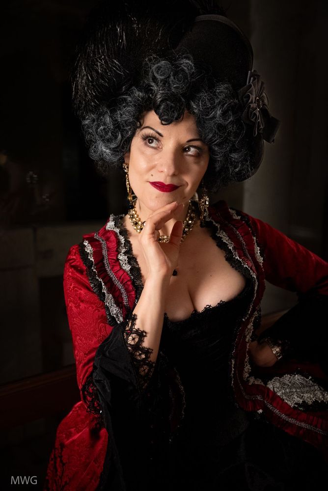 Woman in a red 18th century gown, trimmed in black and silver, looking up and to the right. Clearly contemplating mischief. 