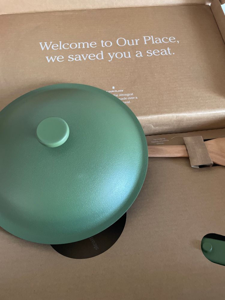 A sage Our Place Always Pan lid in box that reads “welcome to Our Place, we saved you a seat.”