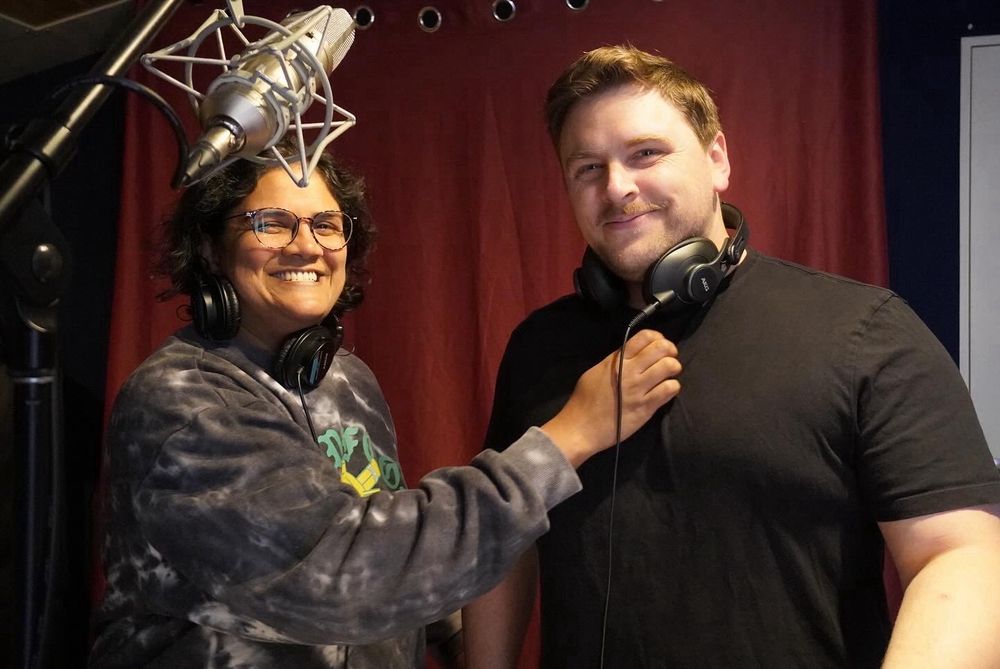 Voice actor Sunanda with headphones around their neck and a big smile on their face, grasps at fellow voice actor Scott Limbrick, also smiling and wearing headphones. There is a silver microphone in the top right corner and a red curtain behind them