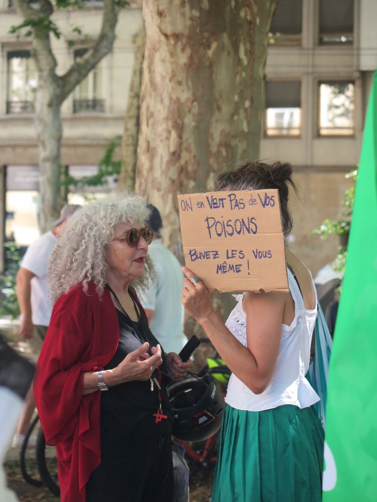 Deux femmes pendant le rassemblement contre la loi Duplomb à Lyon. Une femme aux cheveux blancs avec un casque de vélo à la main et une plus jeune dont le visage est caché par une pancarte disant On ne veut pas de vos poisons, buvez-les vous-même !
