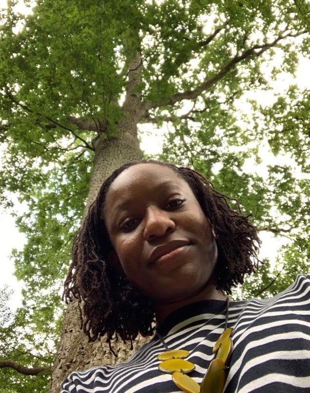 Image shows Dr Victoria Okoye standing underneath a tall tree as she looks down coyly smiling towards the camera at her waist. She wears a black and white striped top, and a necklace which holds a number of yellow-green coloured chunky discs. The sky above is overcast and bright, radiating downward through the tree's leaves and to Victoria.