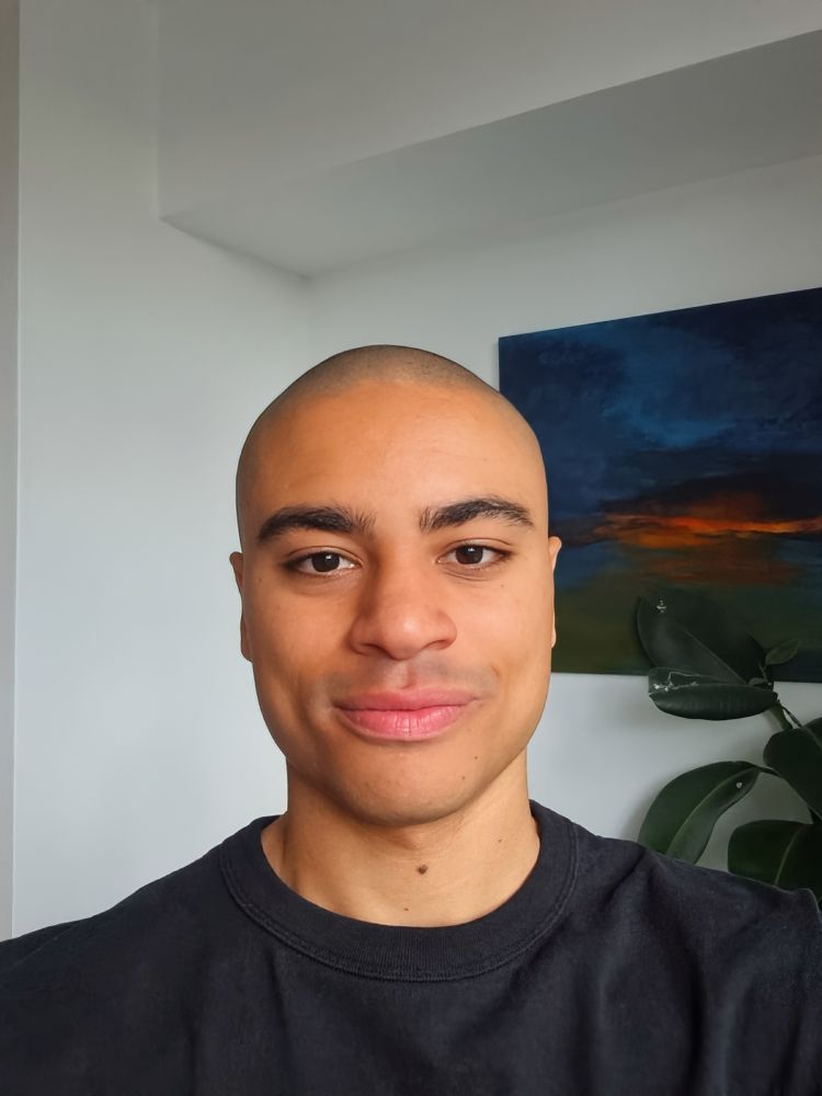 Image shows JMS Scholar, Tyler England, wearing a black crew neck shirt, smiling to the camera in bright daylight. In the background is the upper corner of a white room revealing a tall plant and there is a painting of a rural nightscape on the wall.