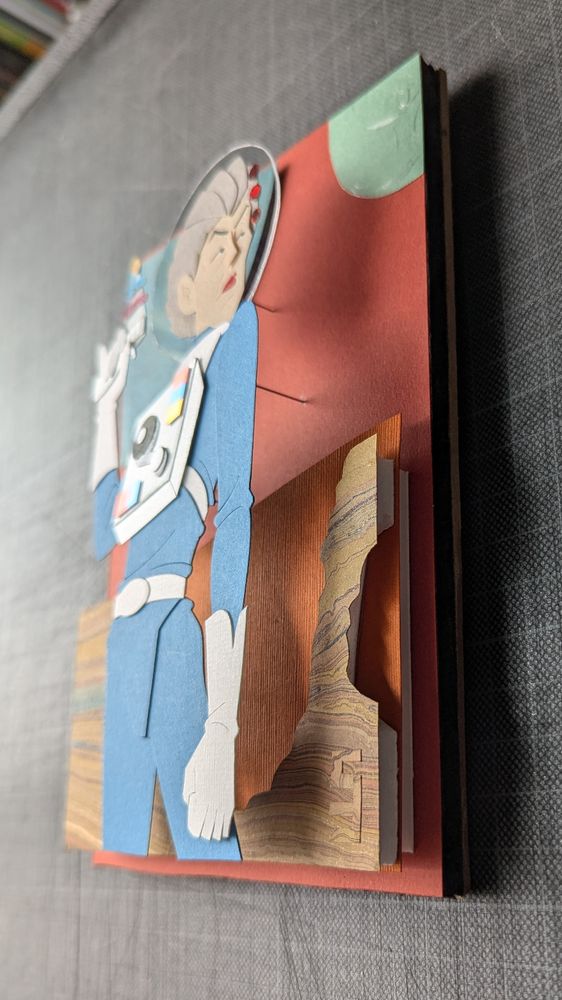 One of three photos of the paper illustration taken from the side to show the height of the piece, with pins, paper, and foam holding up the layers to push the depth.