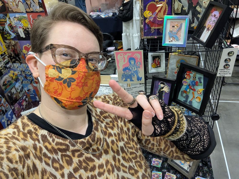 Selfie of the artist giving a peace sign in front of her anime convention booth like a huge dork