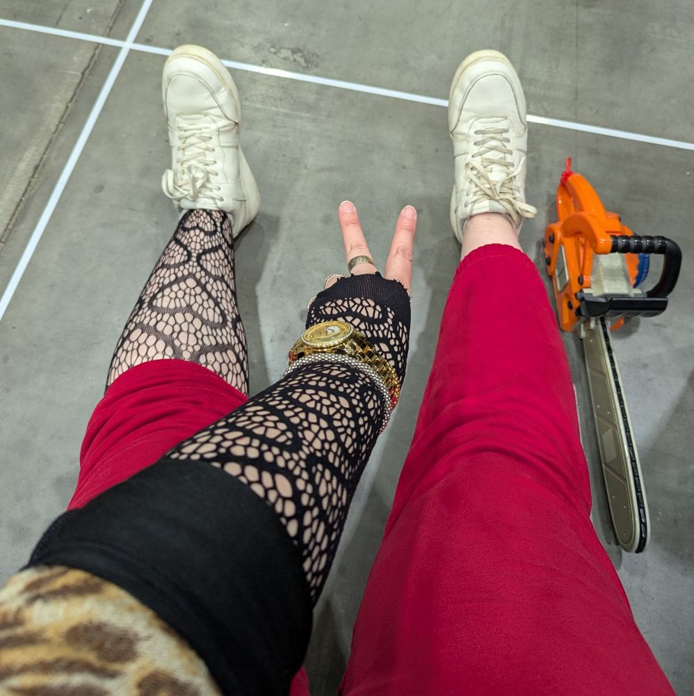 POV photo of the artist's left arm and legs, left arm and leg wearing stockings with a heart pattern on them. There's a chainsaw on the floor even though there's no forest nearby