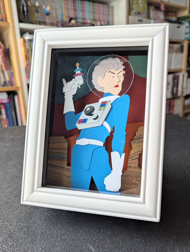 Photo of the illustration, now framed in a white shadow box. It stands on a black surface in front of a bookshelf.