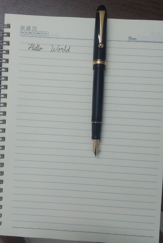 Picture of a Pilot Custom 74 fountain pen on top of a notebook, with "Hello World" written on it.