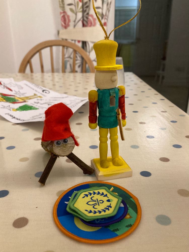 A nutcracker tree decoration standing beside a Spanish present-popping log behind a Chief Scout’s Acorn badge on a pile of other Squirrel Scout badges. 