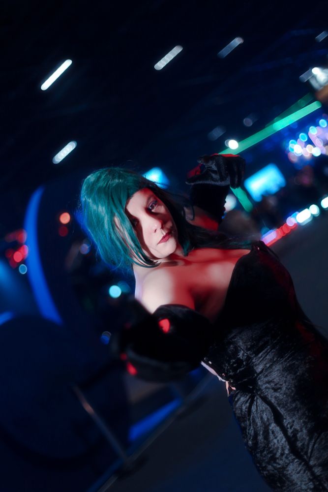 Cosplayer Artemys com a fantasia da personagem Luxúria, do anime Fullmetal Alchemist. A personagem usa um vestido longo, colado ao corpo, na cor preta, com luvas pretas e detalhes em vermelho. A cosplayer usa uma peruca verde musgo.
