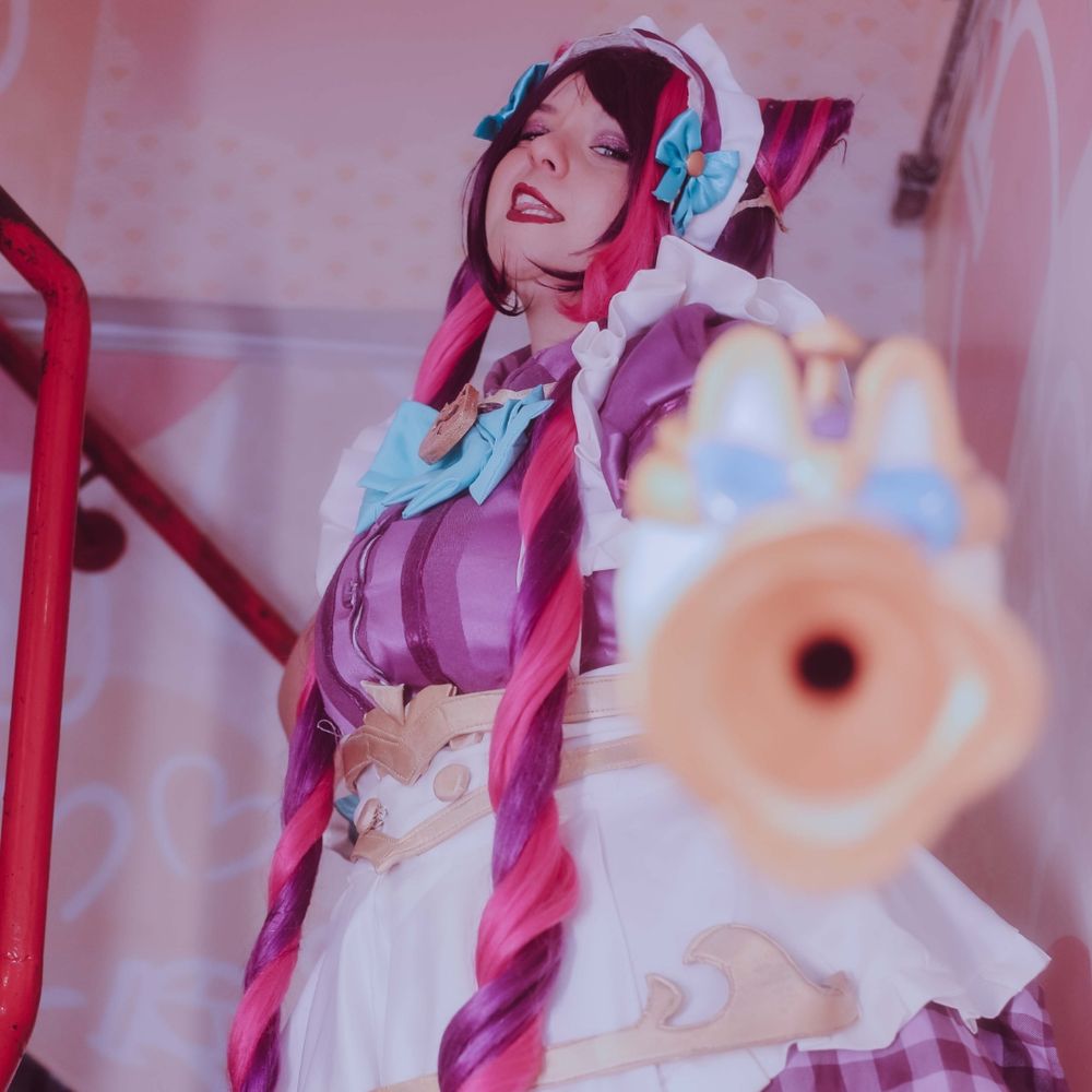 Cosplayer vestida com fantasia da personagem Jinx Kawaii Cafe, do jogo League of Legends. A roupa é um uniforme de 'maid' em tons de roxo, com cabelos em dois rabos de cavalo longos e um avental com babados, e a cosplayer está com uma expressão irreverente, com um dos olhos piscando. Ao fundo, se vê algumas decorações da personagem 'Hello Kitty' na parede.