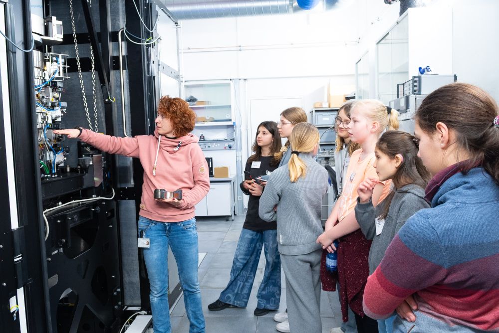 A female scientist explains a large lab instrument to a group of middle school girls during a tour. The students listen closely, surrounded by technical equipment in a lab setting.