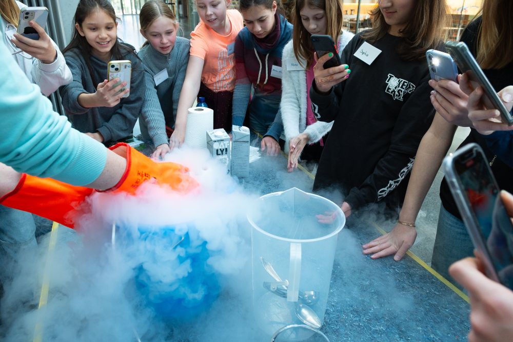 A group of excited middle school students gather around a foggy experiment using liquid nitrogen. A person in orange gloves pours the liquid as the students react with excitement and take photos.