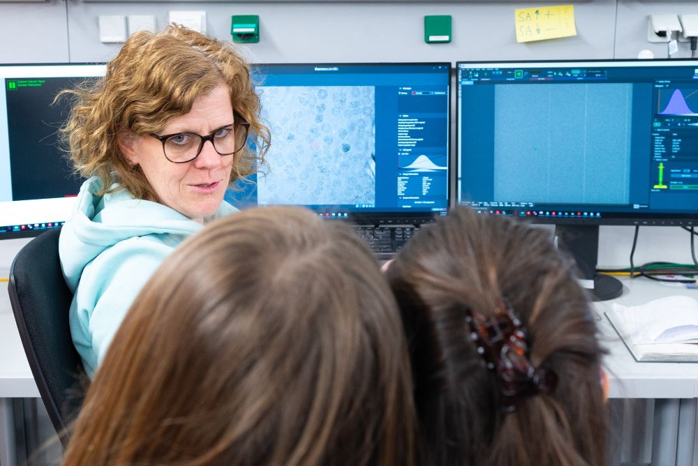 A female scientist wearing glasses sits in front of focused middle school students and explains electron microscopy tomography displayed on a computer monitor.
