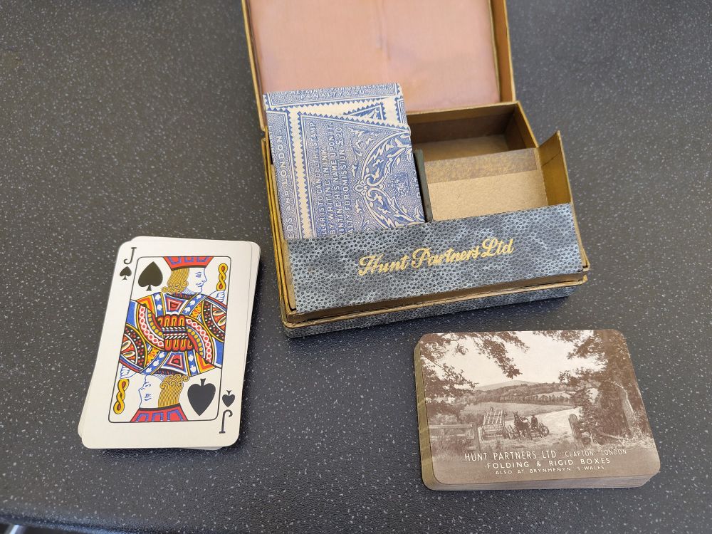 A box that can hold two decks. One deck is displayed with the jack of spades and the face down deck has a photo of an old rural scene. The other deck is wrapped in blue paper