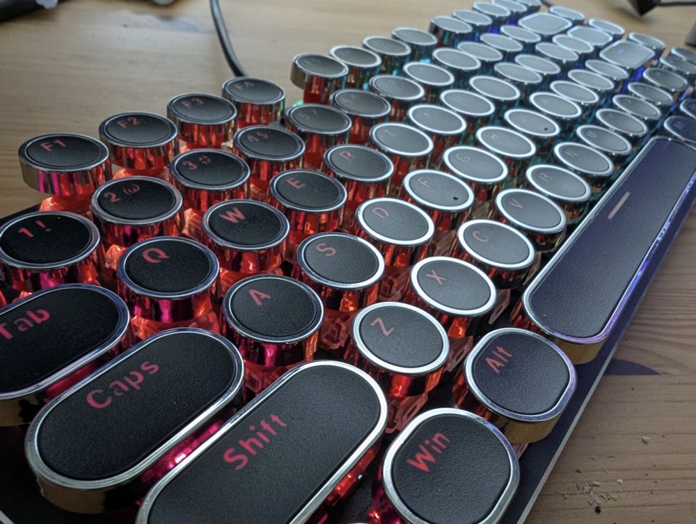 Retro typewriter keycaps on short 84 keyboard. Looking from left down to the right of the keyboard. Each round or rounded key has a black face, chrome rim, and translucent "labels". This picture is with back illumination.
