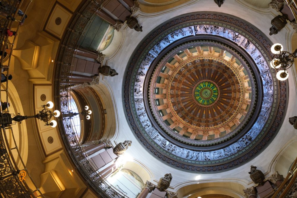 Looking up at the dome of the Springfield Illinois USA capitol building. Part of the railing of the middle and upper floors are visible around and to the left. The dome goes from green, brown, golden, sky blue, alabaster, to golden colors as it descends from topmost to the visible bottom rings.