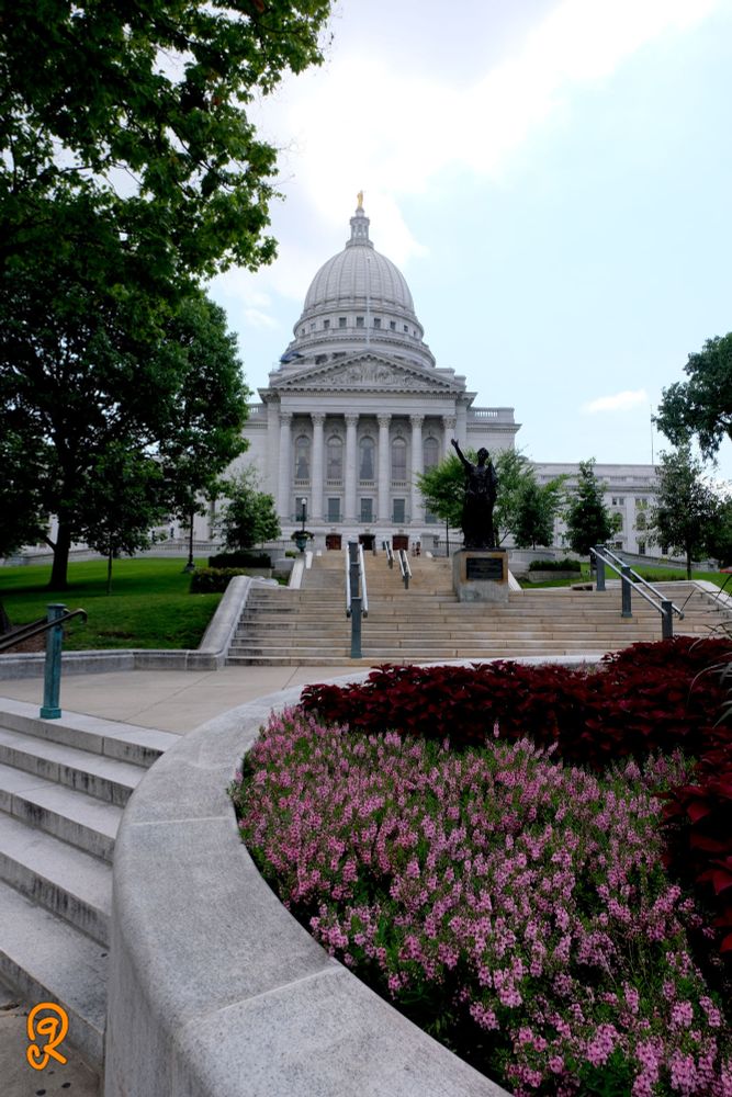 From the bottom steps of the Madison Wisconsin capitol looking at the outside main entry of the building. A large dome with tall columns fronting the entrance. Many steps leading up to the entry. With flowers and trees to welcome you.