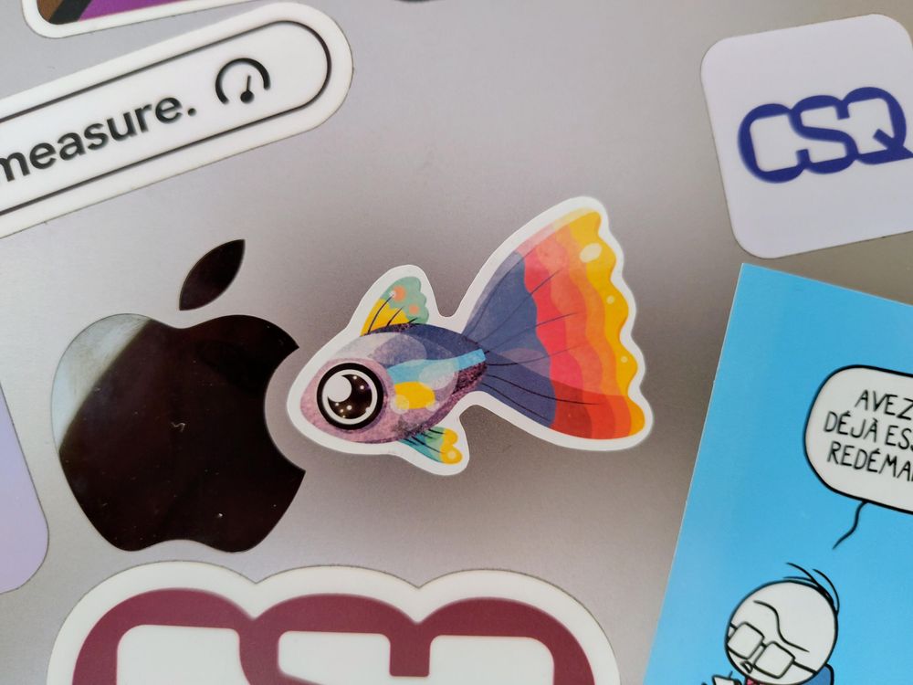A MacBook brushed aluminum surface featuring several stickers. In the center, a colorful little fish.