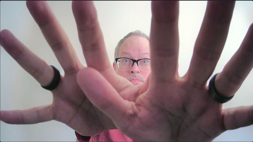 giant splayed hands in front of the camera with a tiny, frightened-looking head with glasses showing between the fingers
