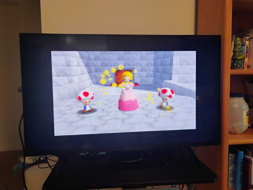 Scene from Super Mario 64 for the Nintendo 64 (N64) via MISTer Pi FPGA on a flat screen. Princess Peach with two Toads on a bridge in front of the castle door.