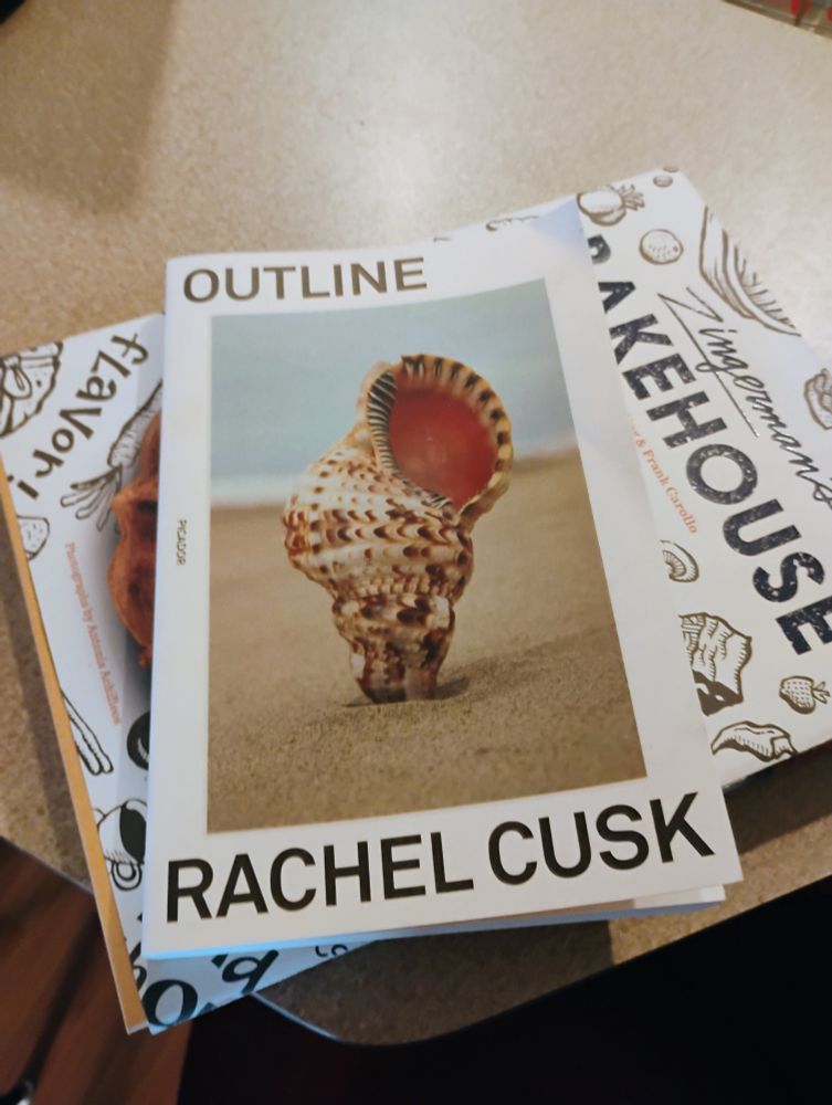 Copy of Outline by Rachel Cusk (with a cover of a seashell planted upright into the sand at the beach) over a copy of the Zingerman's Bakehouse cookbook.